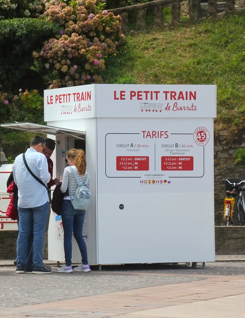 Kleine witte ticketkiosk van Le Petit Train de Biarritz met drie mensen ernaast, omringd door groen en bloemen.