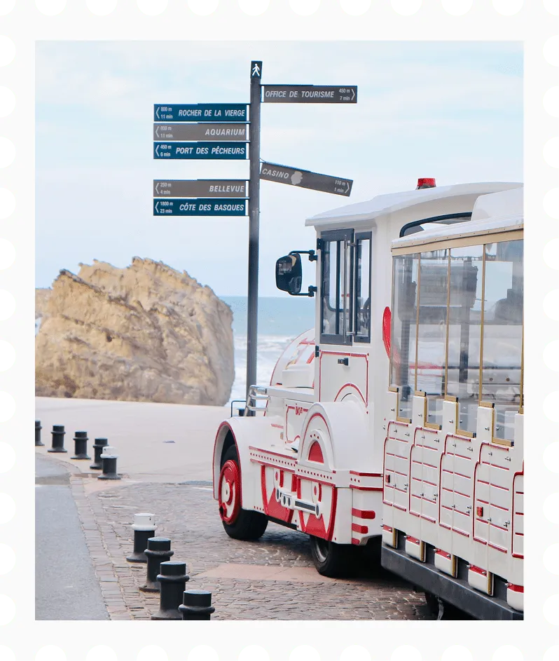 Wit toeristentreintje met rode accenten, geparkeerd bij een uitzichtpunt aan de kust met wegwijzers naar lokale attracties en een grote rots aan zee.