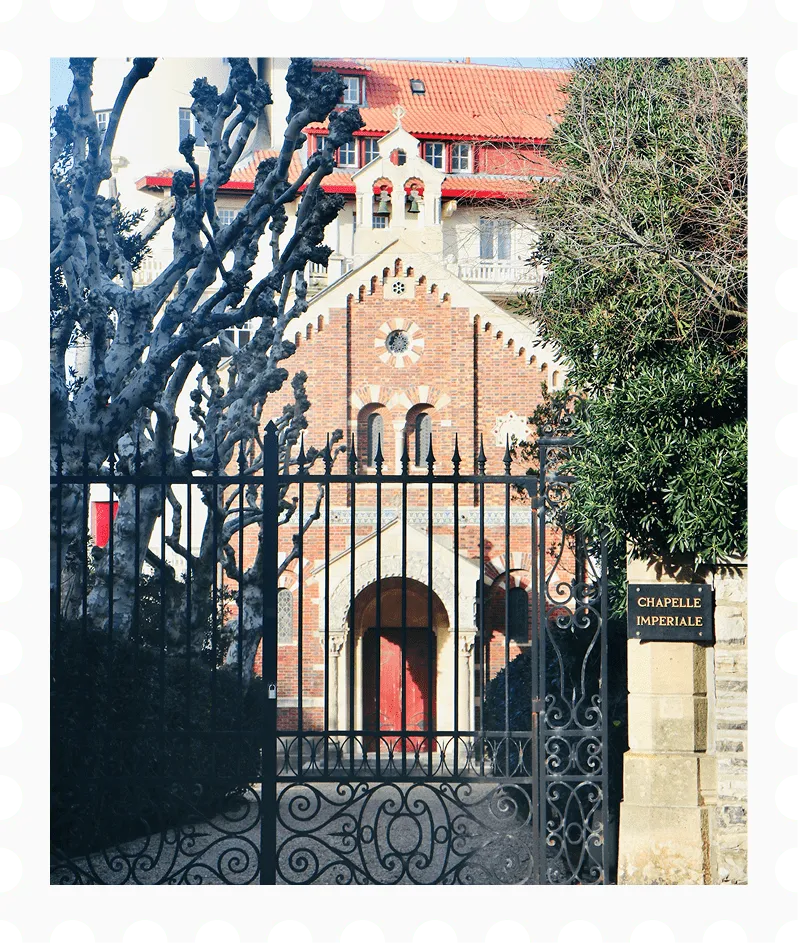 Smeedijzeren hek met fijne motieven voor de Chapelle Impériale, een bakstenen kapel met booggewelfde deur en rode dakpannen.
