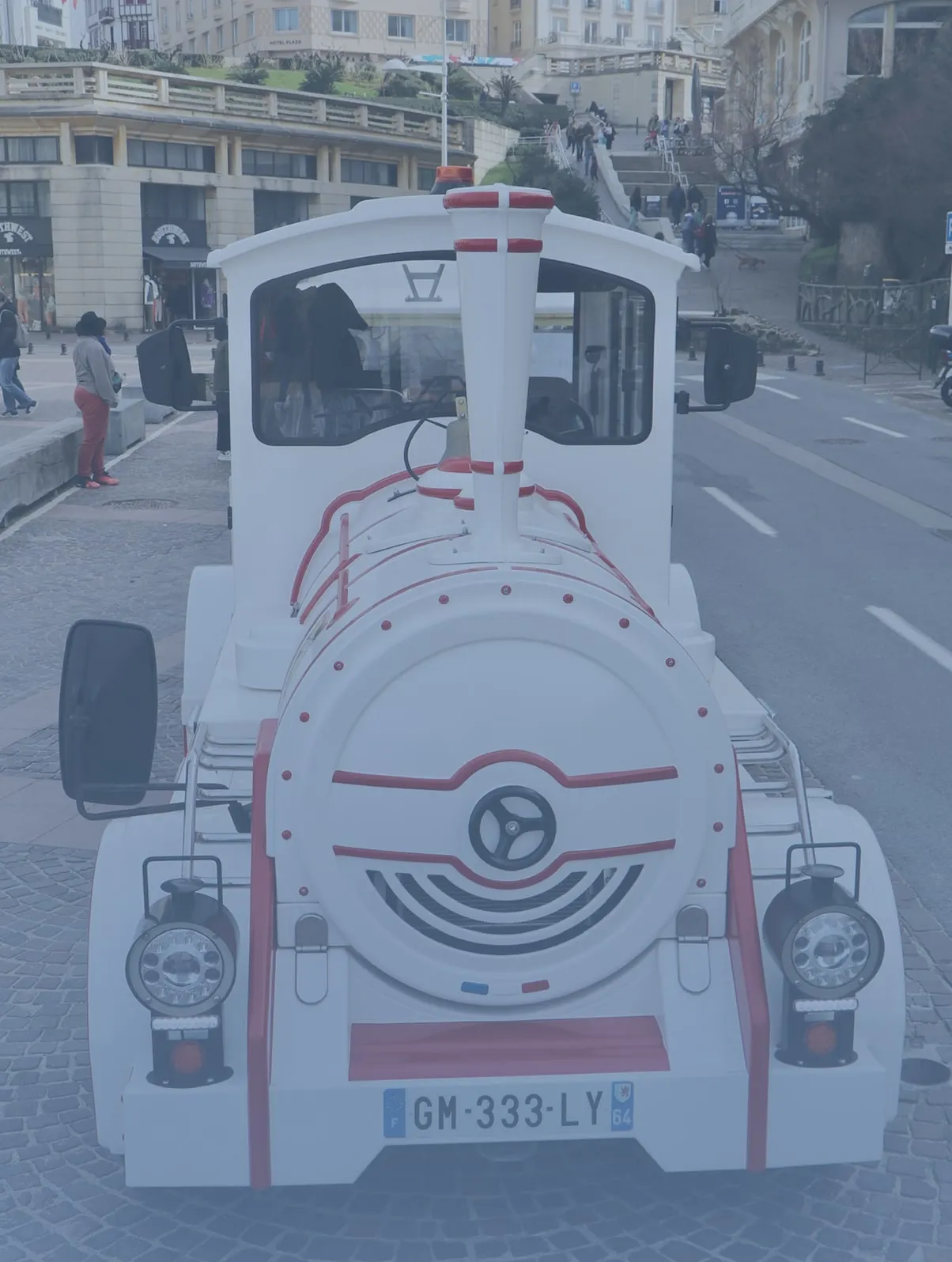 Vooraanzicht van een wit-rood toeristentreintje op een geplaveide straat in een stedelijke omgeving.
