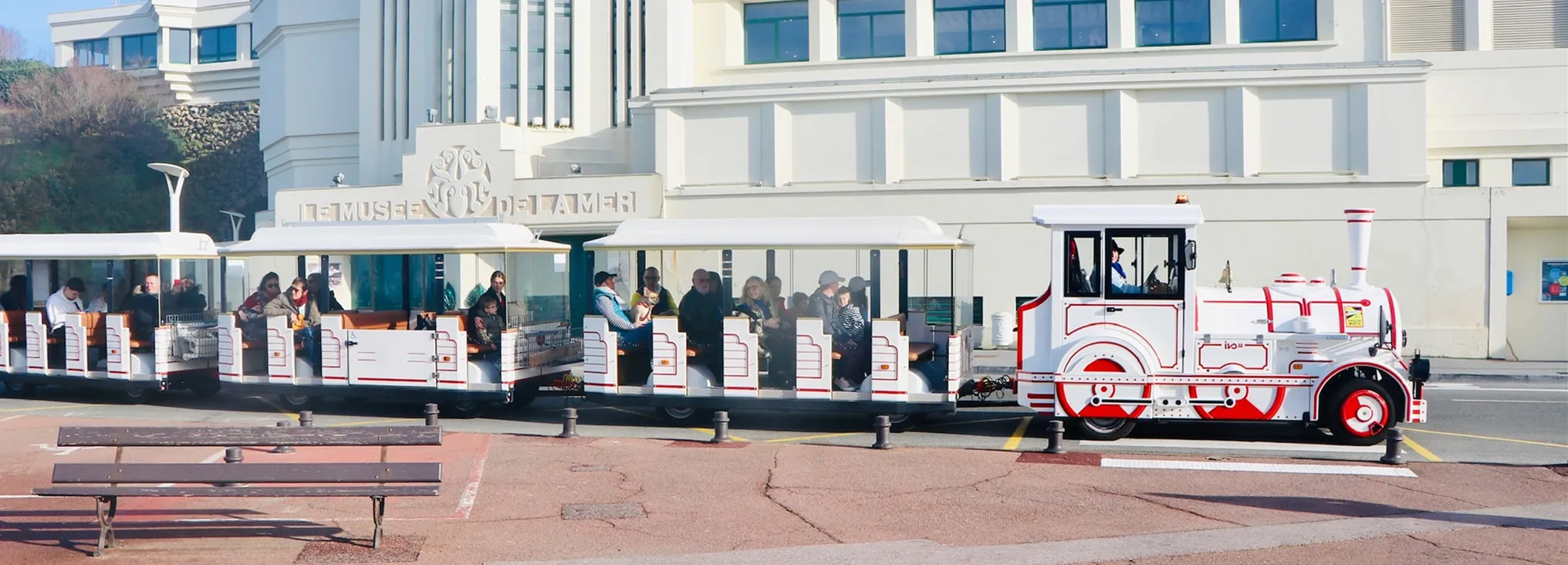 White and red tourist train with open carriages carrying passengers in front of a building labeled Le Musee de l'Ameri.