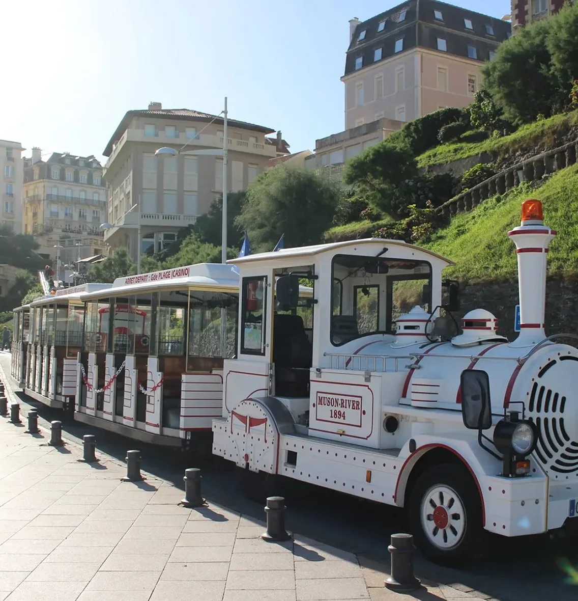 White tourist train with 'Muson River 1894' written on the engine parked on a paved street with buildings and greenery in the background.