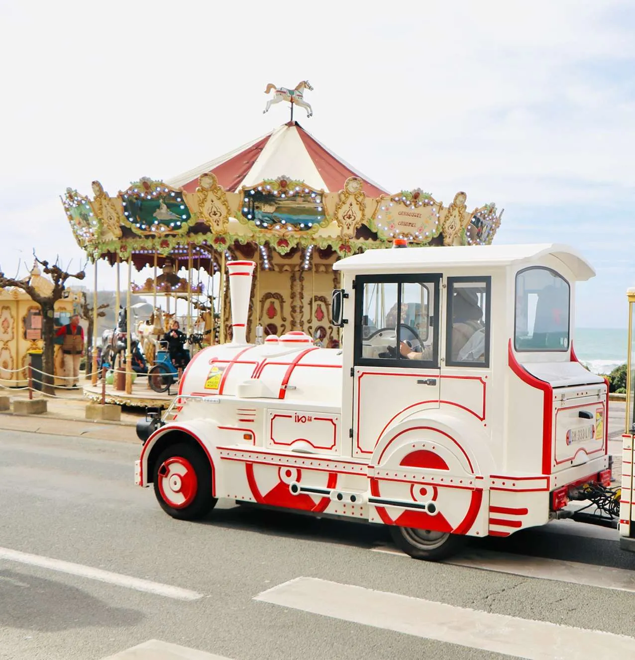 Wit-rood miniatuurtreintje op een weg met een vintage draaimolen en mensen op de achtergrond bij de zee.